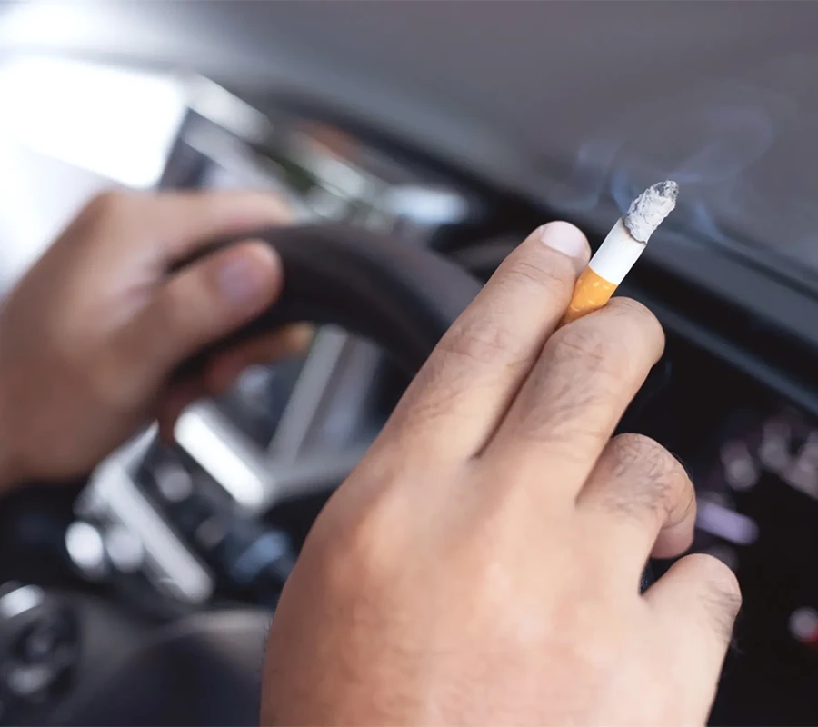Hände einer Person am Lenkrad im Auto, in einer Hand eine angezündete Zigarette.