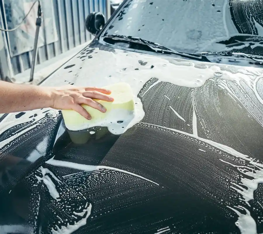 Eine Person führt mit einem gelben Schwamm und Seifenwasser eine Lackpflege-Reinigung an einem schwarzen Auto durch